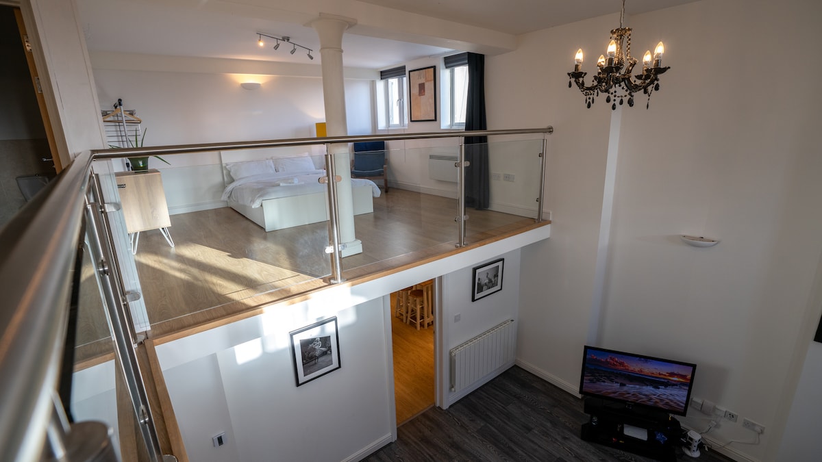 An overview of the open-plan living area and mezzanine bedroom is presented. The stylish space includes double-high ceilings, a television stand, and a framed photo, with natural light illuminating the room. The balcony rail provides a glimpse of the well-sized bedroom above.