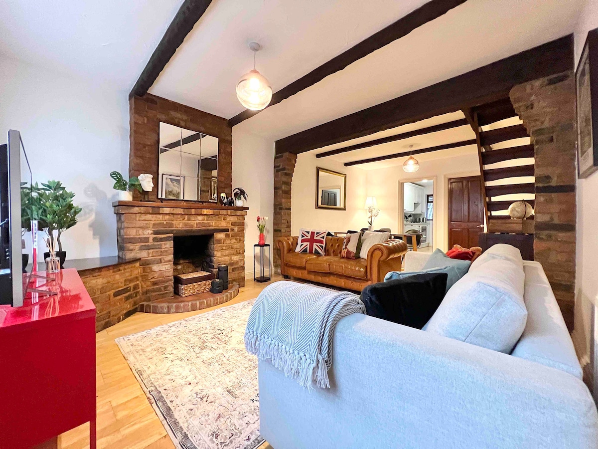 The living area features a cozy seating arrangement with a light-colored sofa and a classic leather couch. A brick fireplace serves as a focal point, complemented by a vintage rug underfoot. Exposed wooden beams and soft lighting enhance the atmosphere of the space.