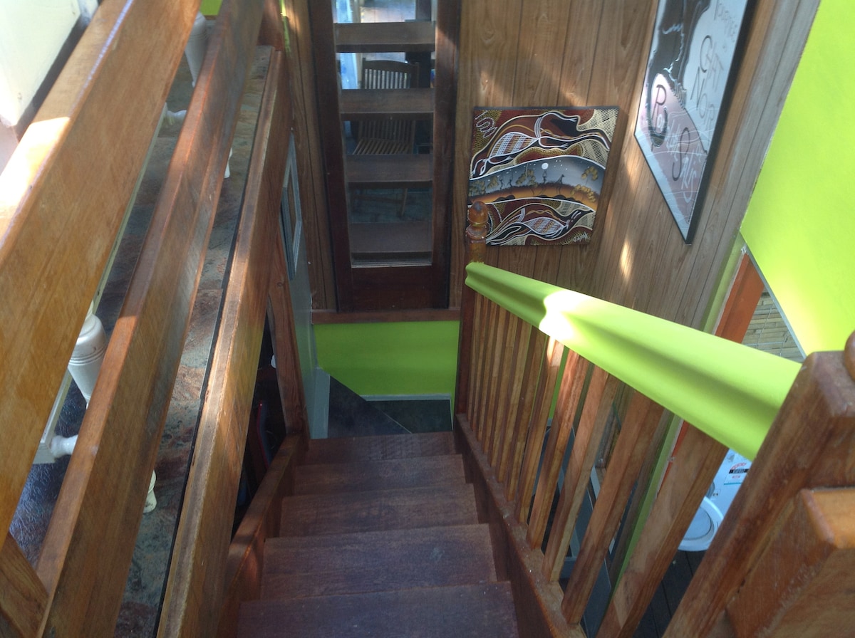 A staircase with wooden steps and a bright green handrail leads down. The surroundings include wooden walls and artwork displayed on the wall, creating a warm and inviting atmosphere. Natural light filters through the nearby windows, enhancing the rustic charm of the space.