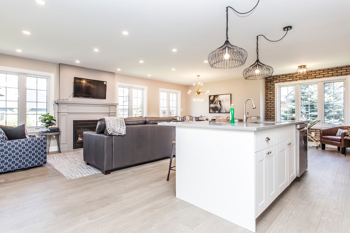 An open-concept living area features a modern kitchen island with seating, complemented by a cozy lounge area. A sleek fireplace is positioned beneath a wall-mounted TV, while large windows allow natural light to fill the space, providing views of the outside landscape.