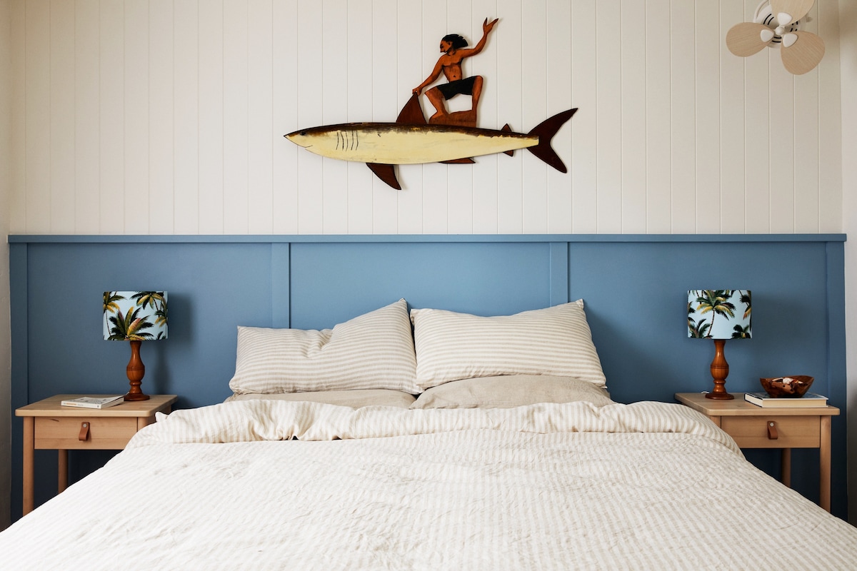 A serene bedroom features a spacious bed adorned with striped linens. On either side of the bed, wooden nightstands hold patterned lamps. A decorative surfboard is mounted above the headboard, complemented by a ceiling fan overhead.