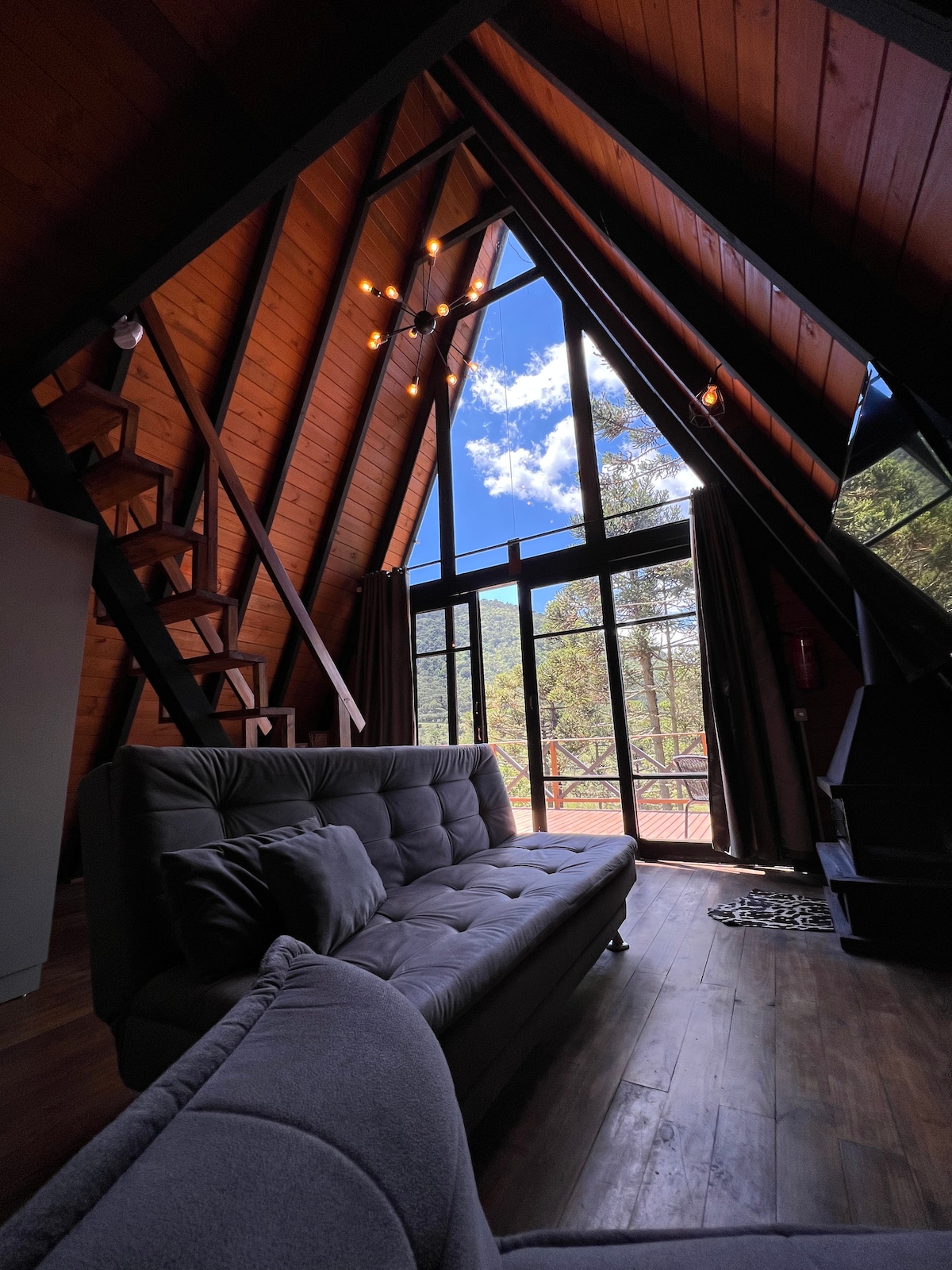 A modern cabin interior features an open design with a high, sloping wooden ceiling. Large glass doors provide access to a scenic outdoor view. Comfortable seating is arranged near the window, with a staircase leading to a lofted area in the background.