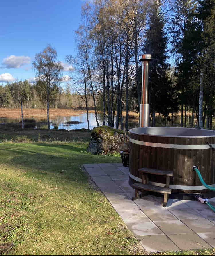 A wooden hot tub is positioned on a stone patio, overlooking a serene water body surrounded by trees. The landscape features a mix of greenery and autumn foliage under a clear blue sky.