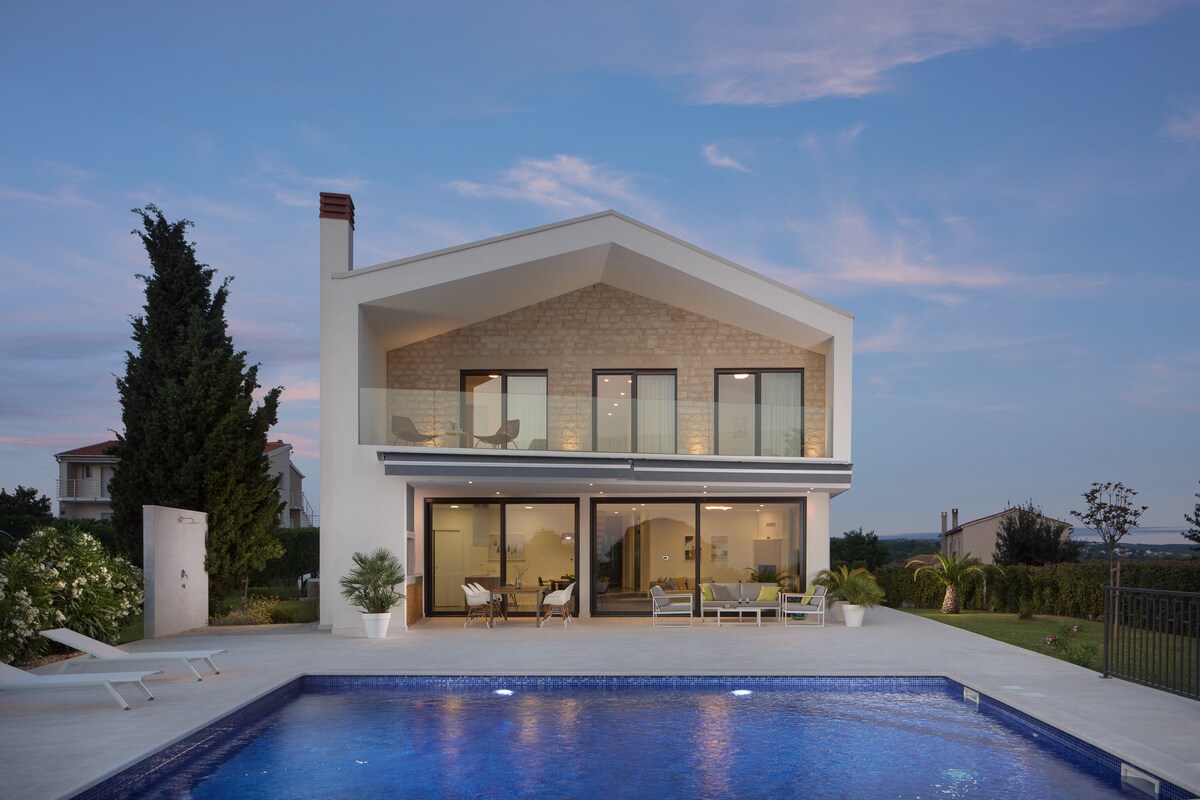 The contemporary villa is showcased at twilight, featuring a spacious terrace that overlooks the illuminated heated pool. Floor-to-ceiling windows allow for visibility into the inviting living area, while surrounding greenery enhances the tranquil atmosphere of the property.