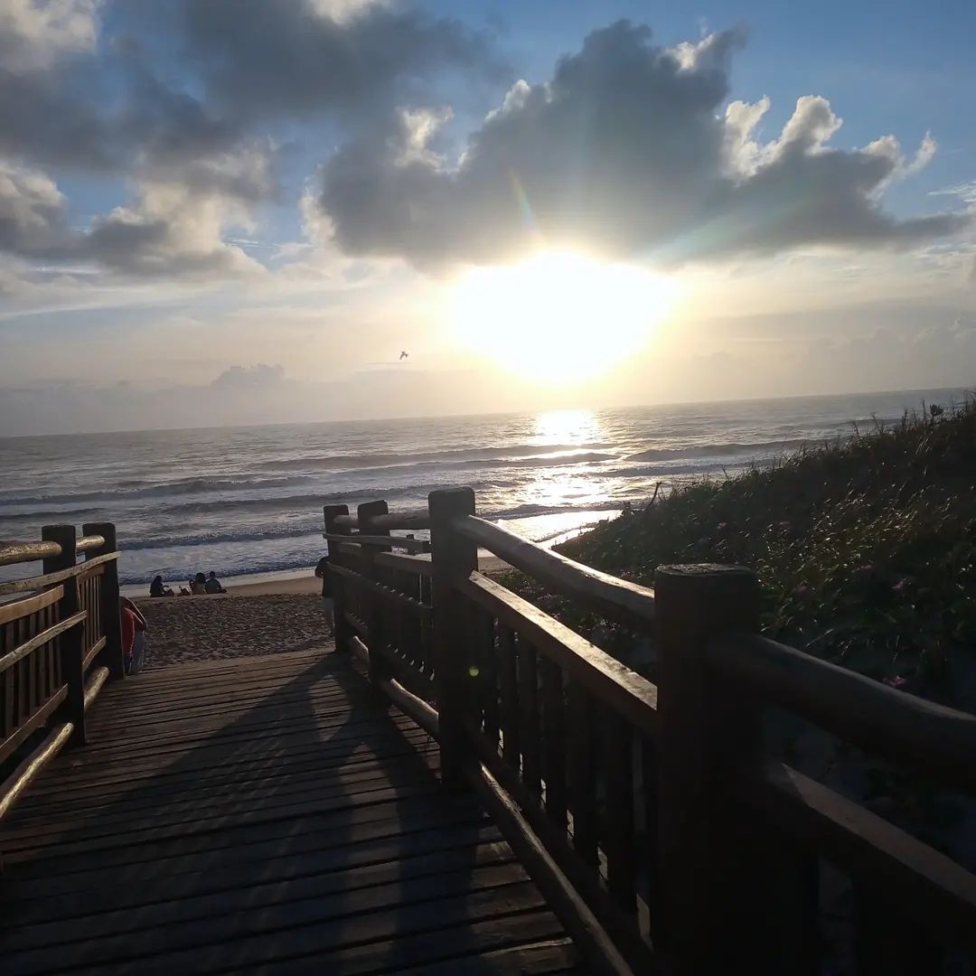 A wooden walkway leads to a beach where the sun is setting over the ocean. The sky is filled with soft clouds, while the glimmering water reflects the warm sunlight. People can be seen sitting on the sand in the distance.