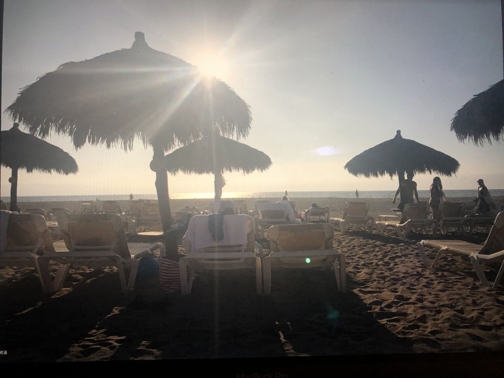 Sunlight filters through thatched umbrellas on a sandy beach. Comfortable lounge chairs are positioned throughout the area, with a view of the ocean in the background. Visitors can be seen walking along the shore, creating a relaxed and inviting beach atmosphere.