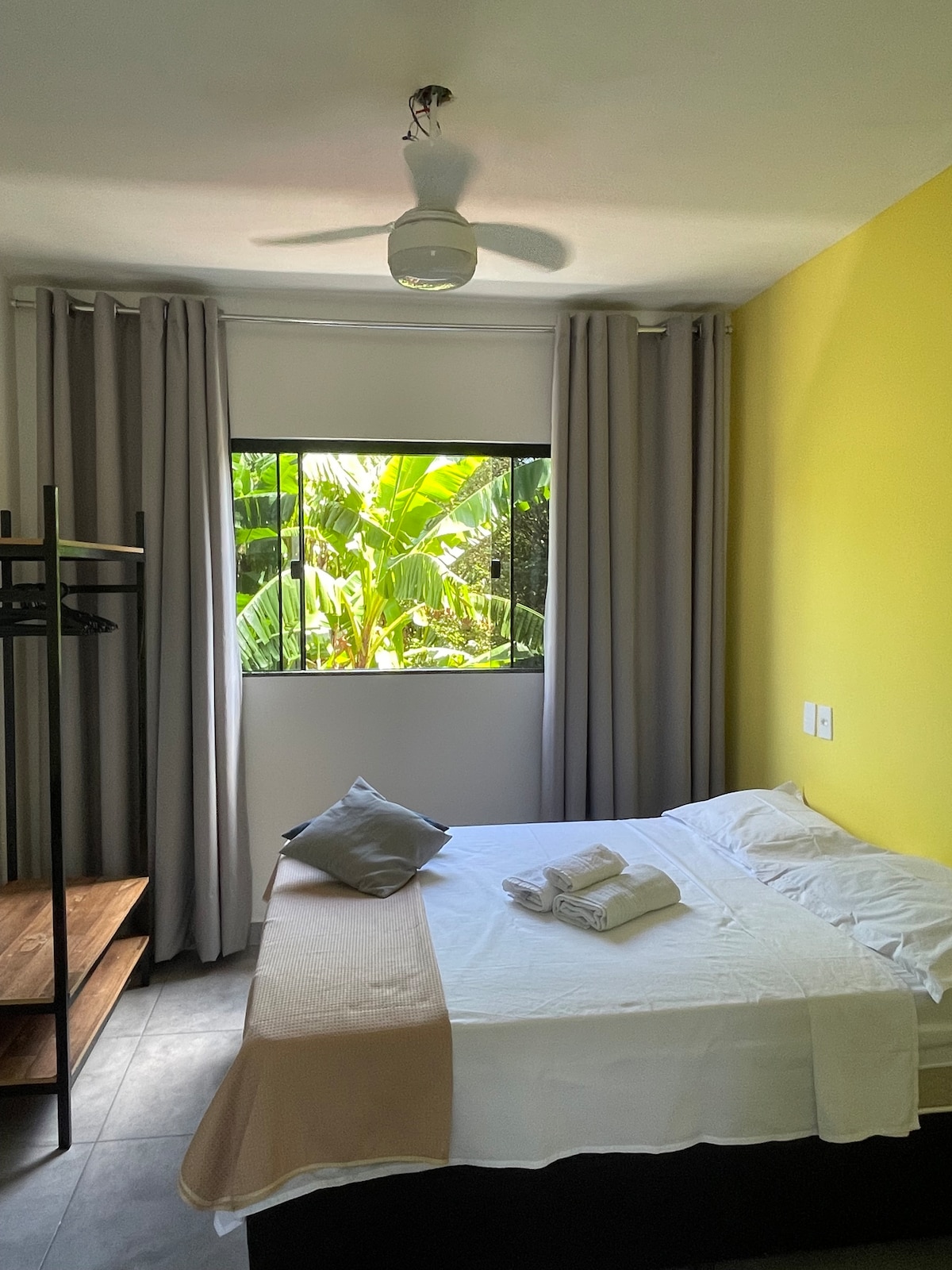 A cozy room features a spacious bed with fresh linens and neatly arranged towels. Natural light filters through a window overlooking lush greenery, while soft gray curtains frame the view. A ceiling fan provides comfort, and a small clothing rack adds practicality.