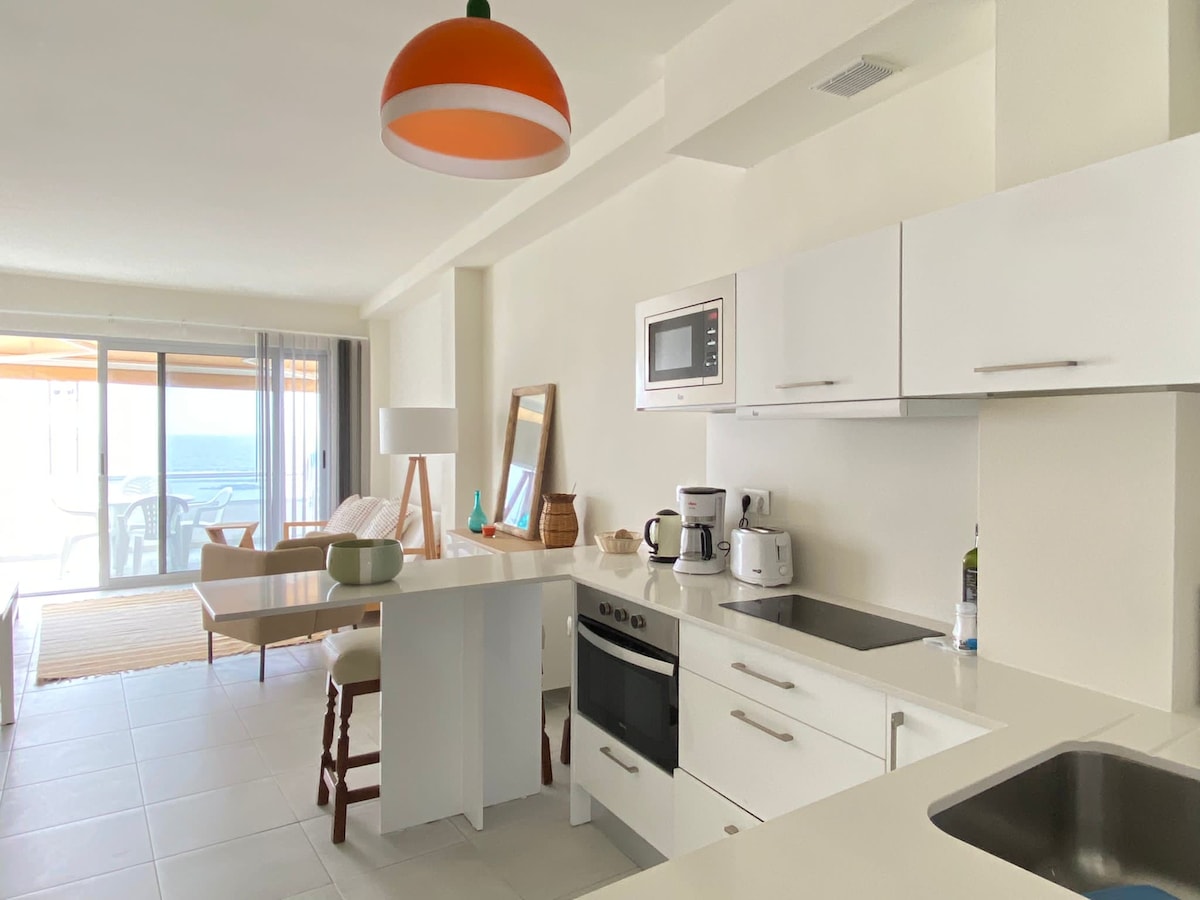 An open kitchen area features modern white cabinetry and appliances, including a stove and microwave. A dining table and chairs are positioned for ocean views, and natural light fills the space through large glass doors leading to the terrace.