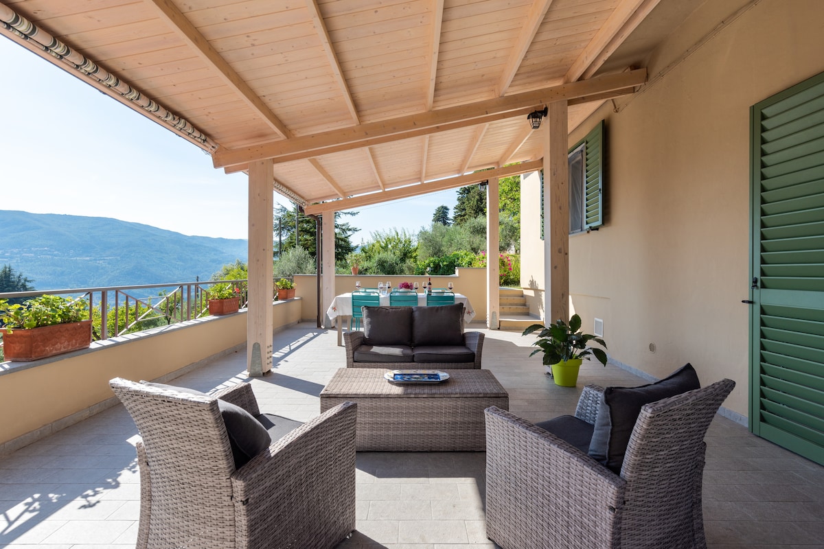 A covered terrace features comfortable seating arrangements with wicker chairs and a coffee table, offering a space for relaxation. Potted plants are placed along the edges, and a view of the surrounding hills is visible in the background under clear skies.