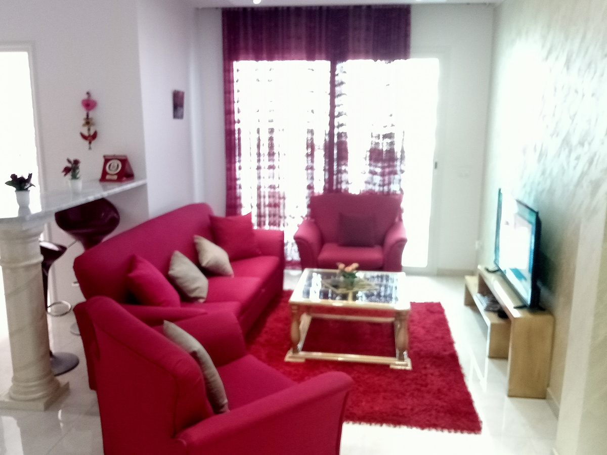 A bright living area features a vibrant red sofa set, including two armchairs and a coffee table. Light-colored walls complement the dark curtains, allowing soft light to filter through. A plush red rug adds warmth to the space, enhancing the atmosphere of comfort.