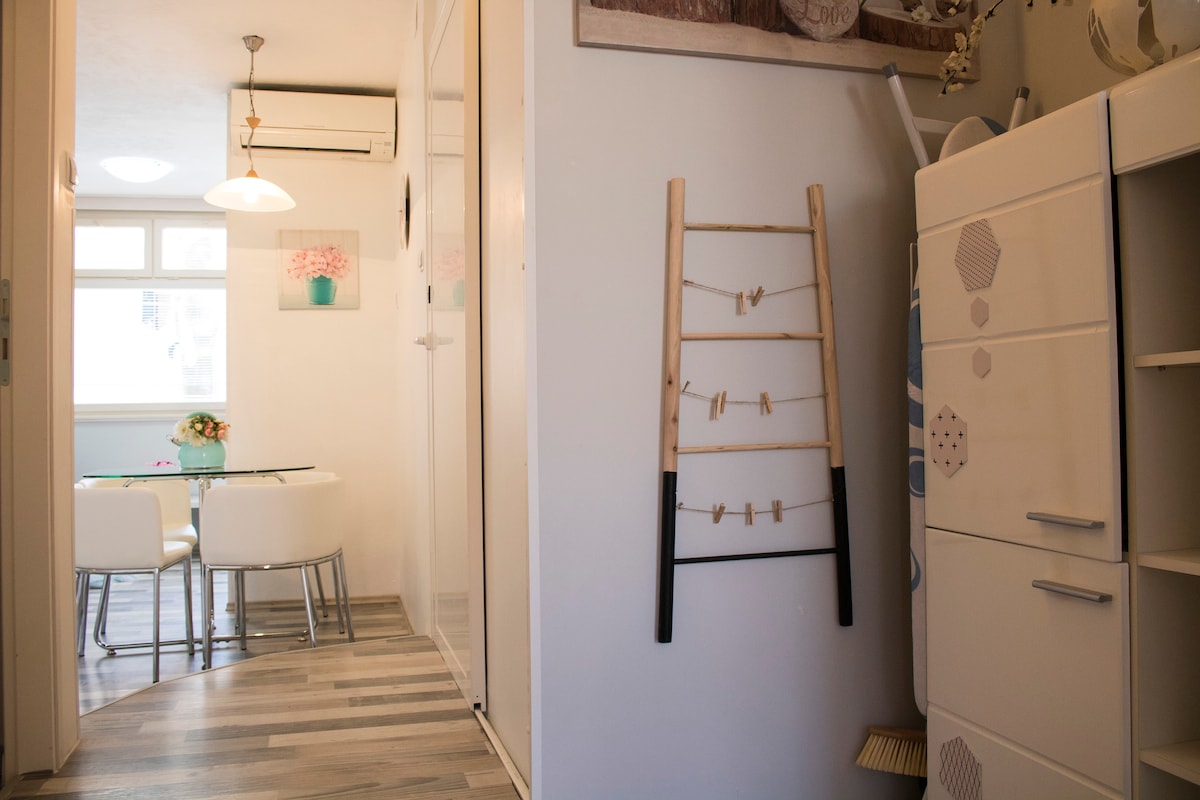 A welcoming entryway is depicted, featuring a wooden ladder for hanging items and a white cabinet with a textured design. Light wooden flooring complements the space, while a dining area is visible in the background, illuminated by natural light filtering through the window.