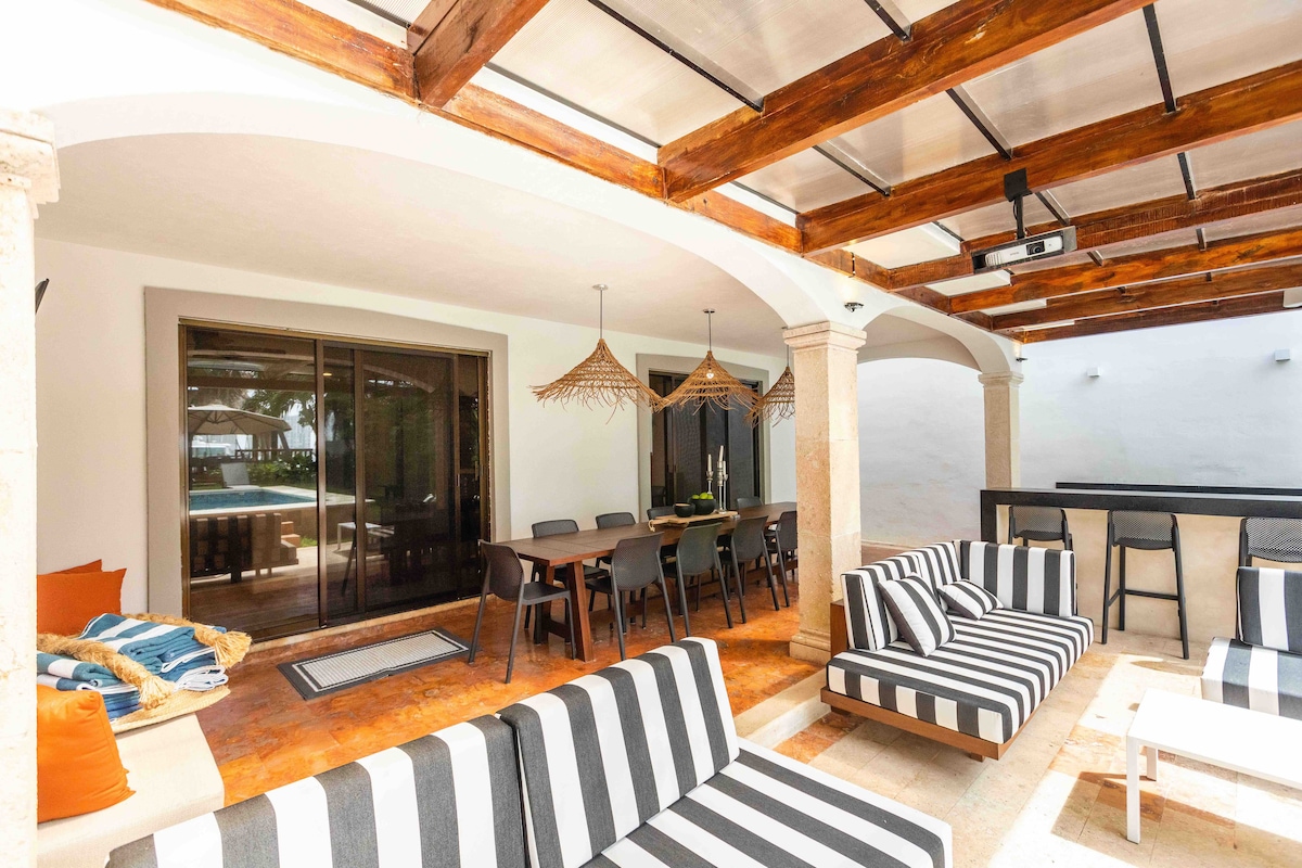 The outdoor patio area features a shaded seating arrangement with striped lounge chairs and a wooden table. A dining space with stylish chairs is positioned nearby, offering views of the pool area through large glass doors, enhancing the flow between indoor and outdoor spaces.