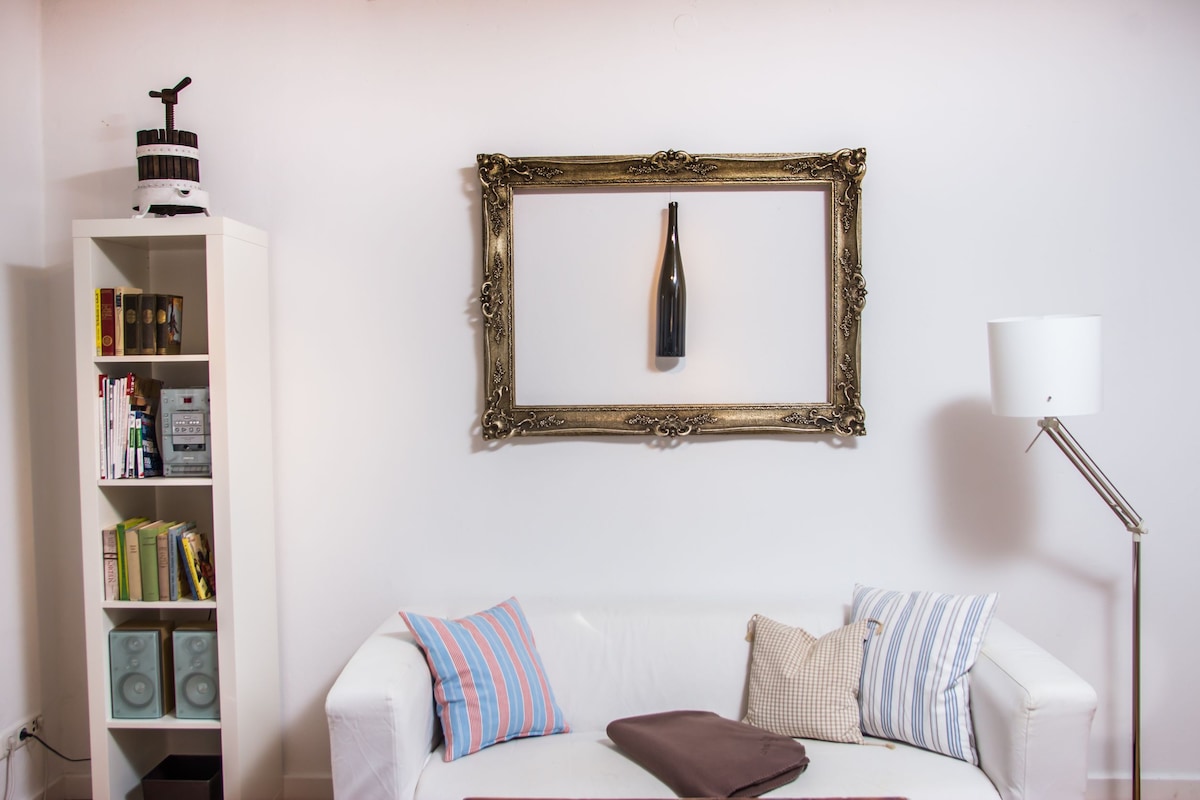 A white sofa is positioned against a light-colored wall, accented by decorative pillows. Above, a framed wall decor piece holds a bottle, adding a touch of character. A sleek lamp stands nearby, and a bookshelf displays a variety of books and decorative items.