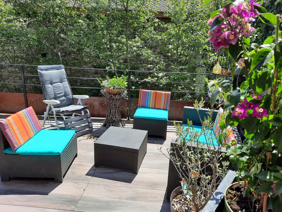 A private terrace features comfortable seating with bright cushions, alongside a small table. Lush greenery adds to the tranquil atmosphere, while blooming flowers provide colorful accents. The surrounding area offers a peaceful setting with no visible neighbors.