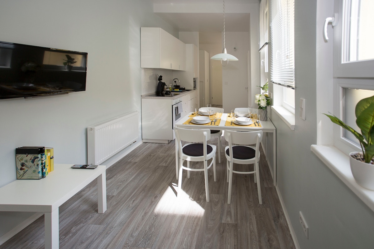 A modern kitchenette is complemented by a white dining table set for four, featuring simple light-colored chairs. A flat-screen TV is mounted on a light wall, while natural light pours in through multiple windows, creating an open and bright ambiance.