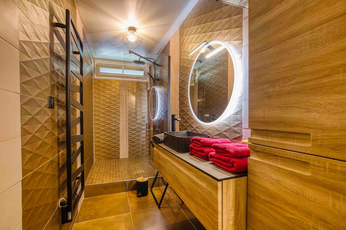 The bathroom features a spacious walk-in shower with a modern design, accented by textured tiles. A large illuminated mirror is mounted above a wooden vanity, complemented by neatly stacked red towels. Additional wooden cabinetry provides storage, contributing to an organized and inviting space.