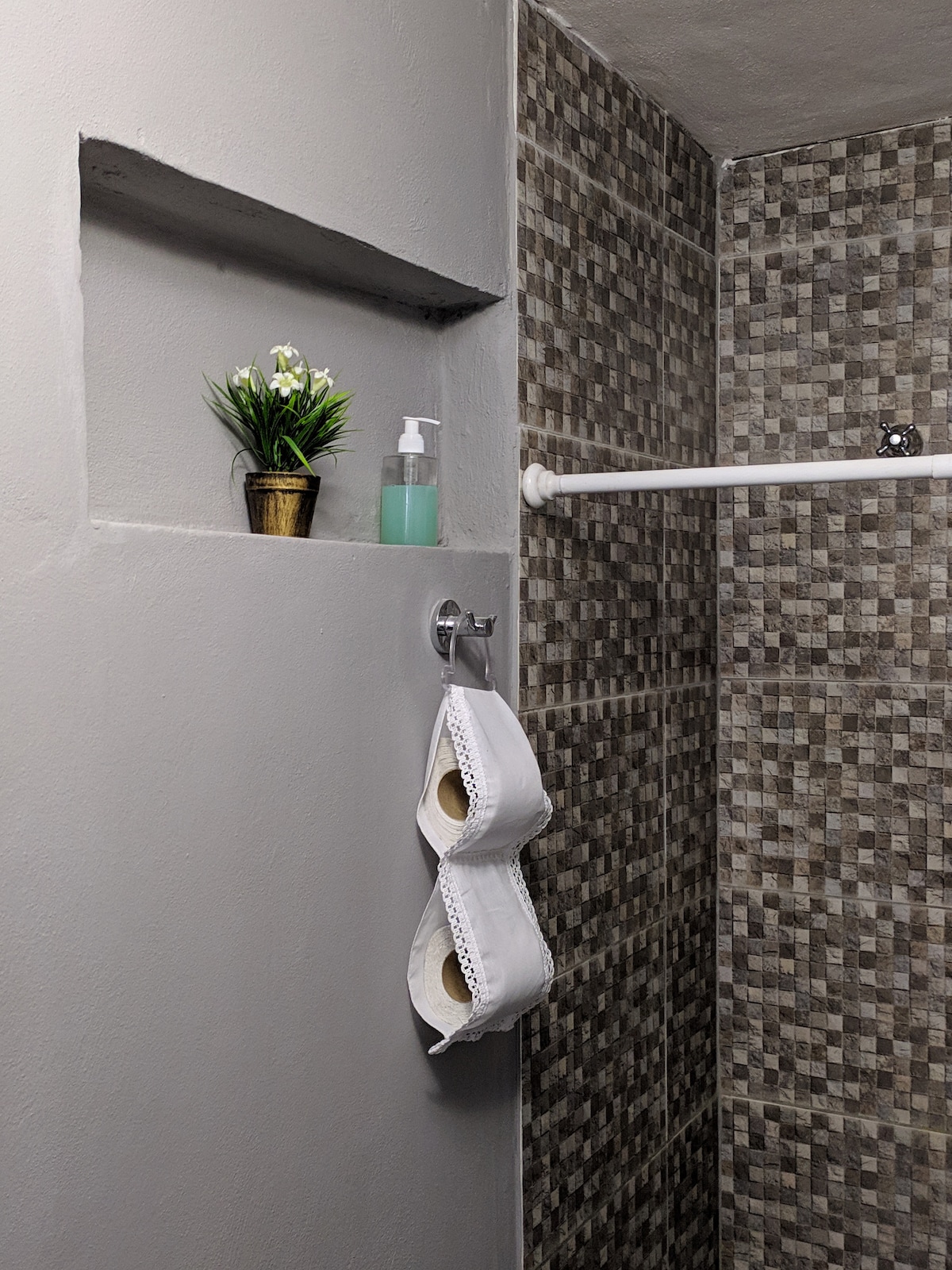 The bathroom features a tiled wall with a decorative niche holding a small potted plant and a soap dispenser. Two neatly rolled towels are displayed on a towel rack, enhancing the practical design of the space.
