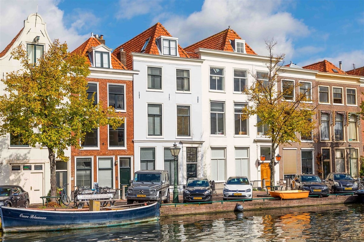 A scenic view of a charming canal-front building featuring a mix of architectural styles. Large windows punctuate the façade, providing ample natural light. Trees line the canal, and various boats and cars are visible along the waterside, creating a lively urban atmosphere.