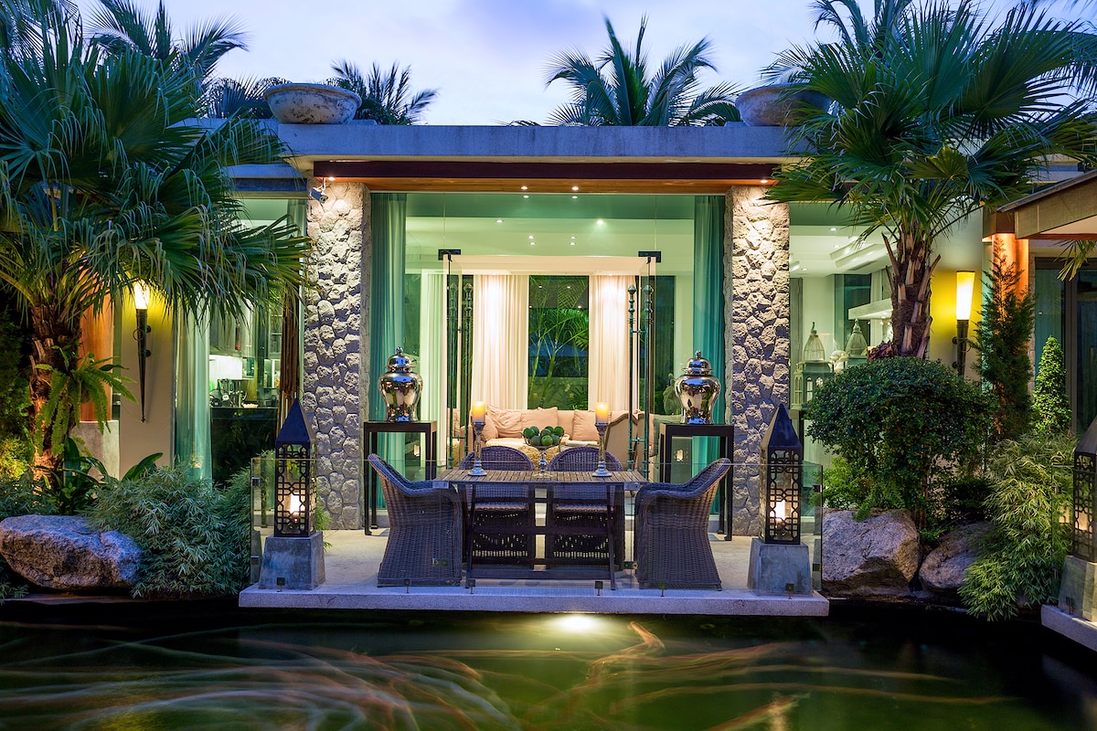 An outdoor living area is framed by lush tropical plants, with a seating arrangement centered on a stone patio. Lanterns provide ambient lighting, reflecting softly on the water surface of the Koi carp pond. Large glass doors lead to the interior, enhancing the connection with nature.