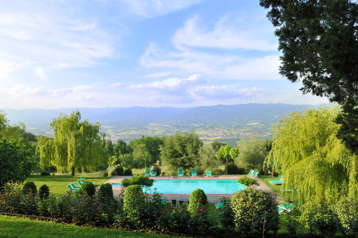 A spacious outdoor area is featured, showcasing a seasonal swimming pool surrounded by well-maintained greenery. Sun loungers are arranged around the pool, while trees and bushes add to the serene landscape. Scenic hills and valleys extend in the background, under a clear blue sky.