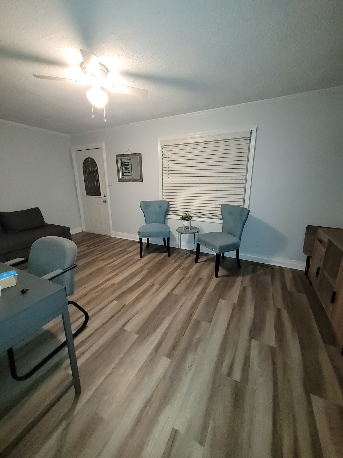 A well-lit living area features light wood flooring and a neutral color palette. Two blue chairs are positioned beside a small table with a plant. A comfortable sofa is located against one wall, while a door and window allow natural light into the space.
