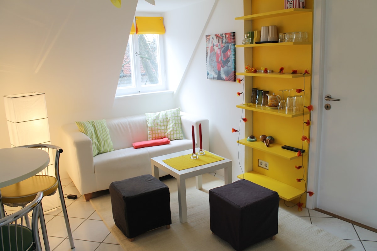 A bright living area features a minimalist white sofa adorned with green cushions. A small table is set with candles, and colorful shelving displays glassware and decor. Natural light filters through a window, illuminating the space, while a light rug adds warmth.
