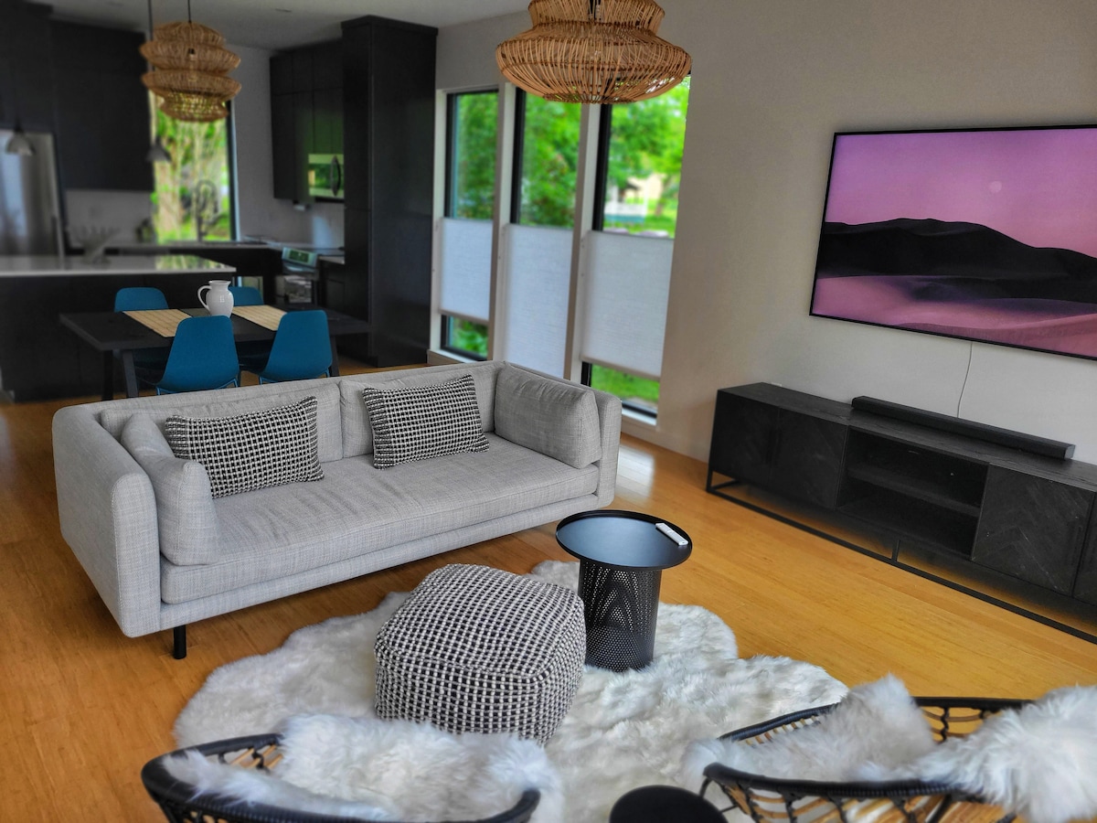 A modern living area showcases a light gray sofa paired with a textured pouf on a soft white rug. A flat-screen TV hangs on the wall, while a dining table with blue chairs is visible in the background. Large windows allow natural light to fill the space.