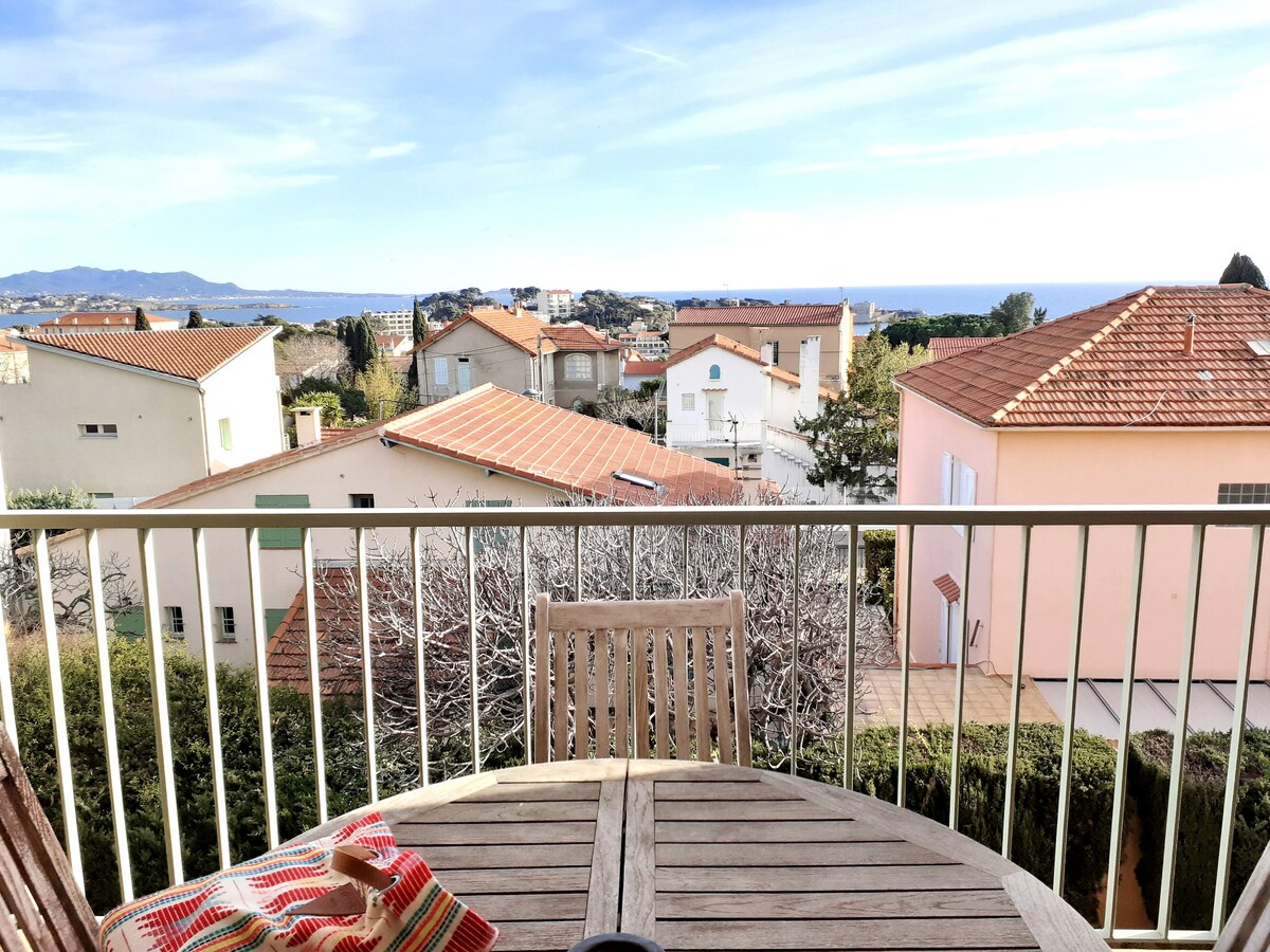 A wooden table and chairs are positioned on a balcony, offering a panoramic view of the surrounding rooftops and the sea in the distance. The sunlit landscape features a clear sky and the gentle curve of hills, creating a serene setting for relaxation.