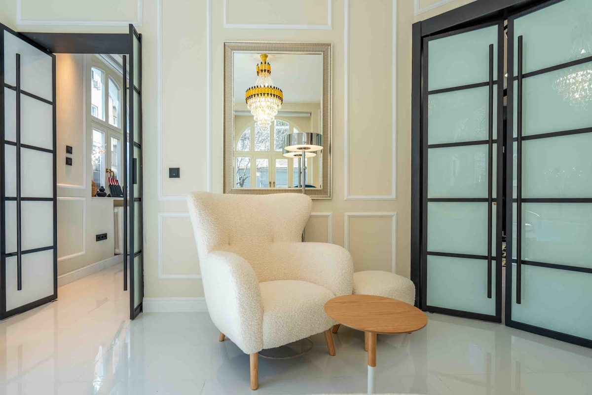 A modern living area is showcased, featuring a plush, white armchair paired with a small wooden side table. Elegant doors with frosted glass panels are visible in the background, along with a mirror reflecting a stylish chandelier above.