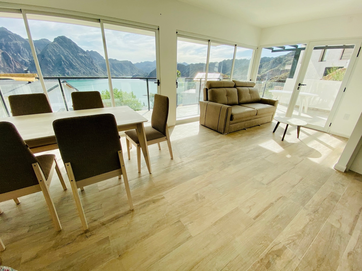 The bright living area features large sliding glass doors, offering views of the surrounding mountains and lake. A spacious dining table is accompanied by four chairs, while a comfortable sofa is situated nearby. Natural light fills the space, highlighting the warm wood flooring.