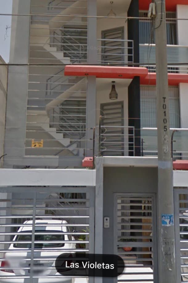 The exterior of a modern multi-story building is depicted, showcasing a clean facade with a combination of red and gray accents. A secure entrance is visible, along with metal railings leading to the upper floors. A staircase is clearly outlined, providing access to the apartments.
