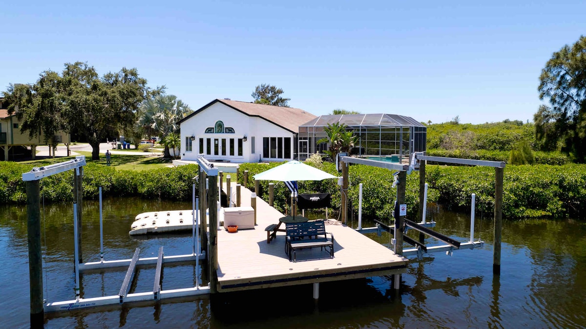 A spacious private dock extends into the Braden River, featuring a shaded seating area and lounge chairs. The view includes the home's rear, showcasing large windows and a screened-in pool area surrounded by lush greenery.