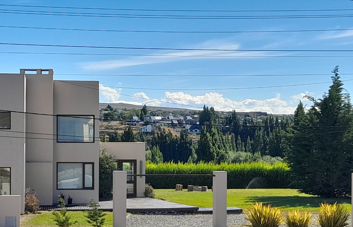 The modern structure is set against a vast sky and rolling hills. Lush greenery surrounds the property, with distant houses visible on the hillside. The landscaping features neatly arranged shrubs and a gravel pathway leading to the entrance.