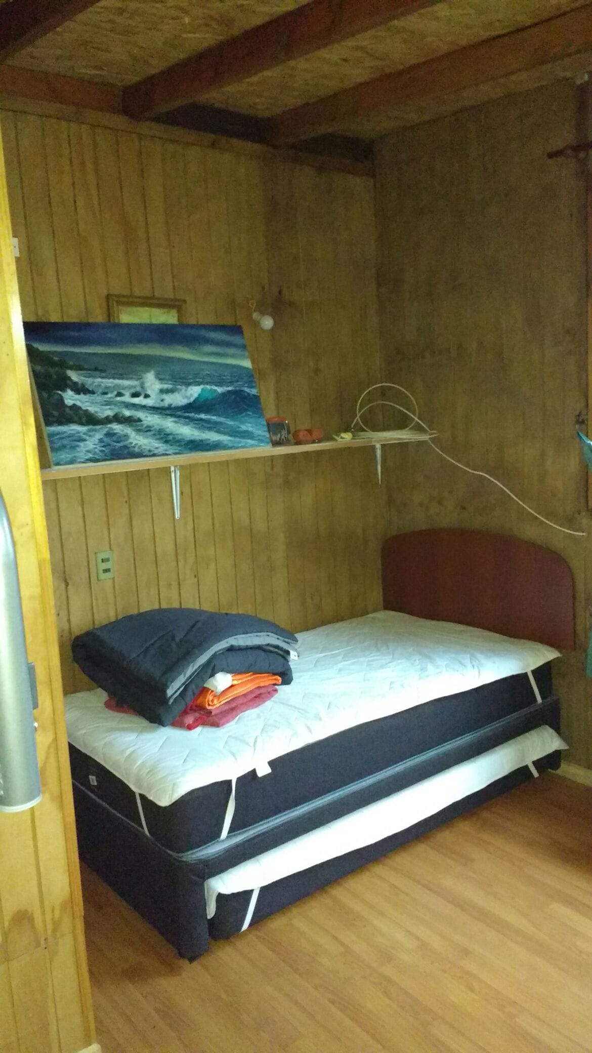 A small sleeping area features a single bed with a layered blanket and neatly folded towels. A wooden shelf holds a painting of ocean waves, while a simple chair is positioned next to the bed. The room is illuminated by natural light from an adjacent window.