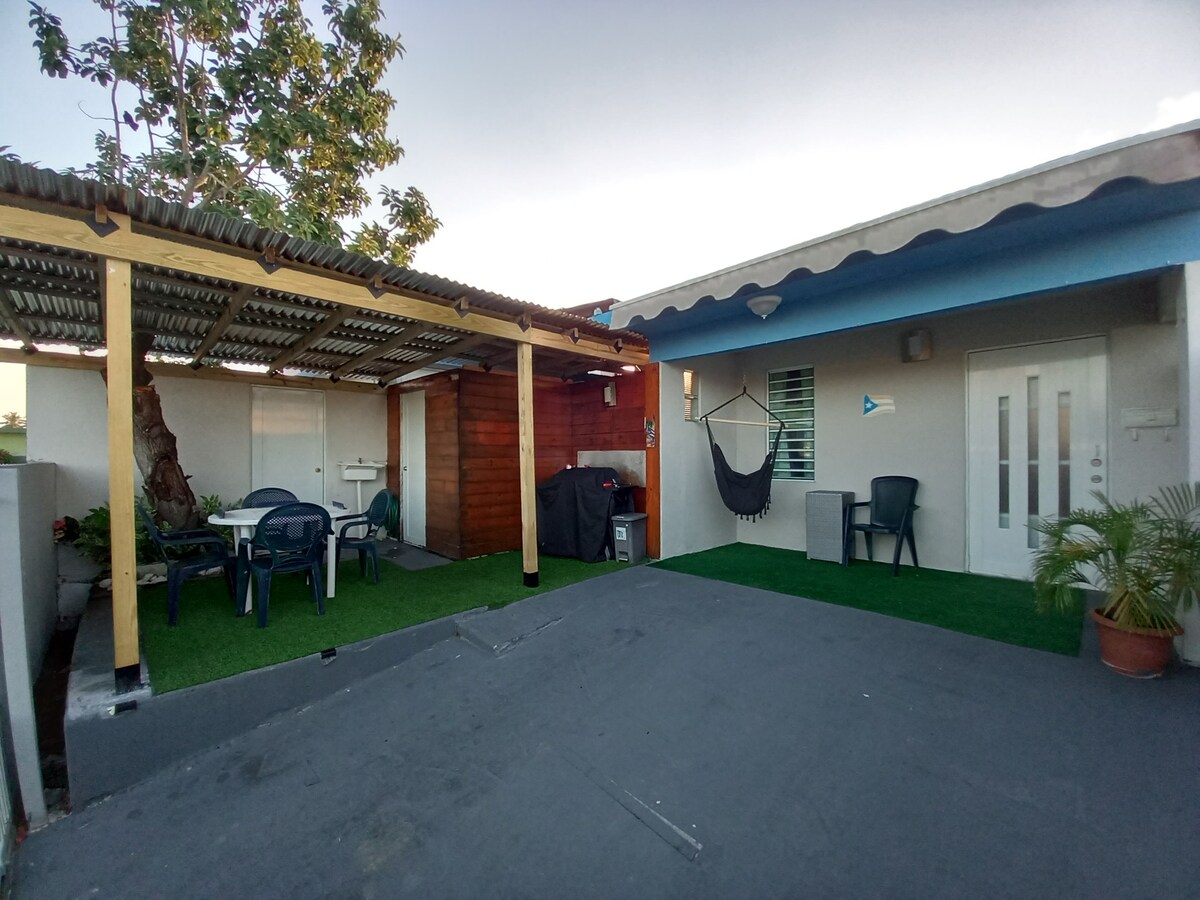 A rooftop space features a covered seating area with two chairs and a small table, surrounded by greenery. A hammock is suspended nearby, offering a relaxed atmosphere. The area is finished with artificial grass, and a wooden structure adds rustic charm.