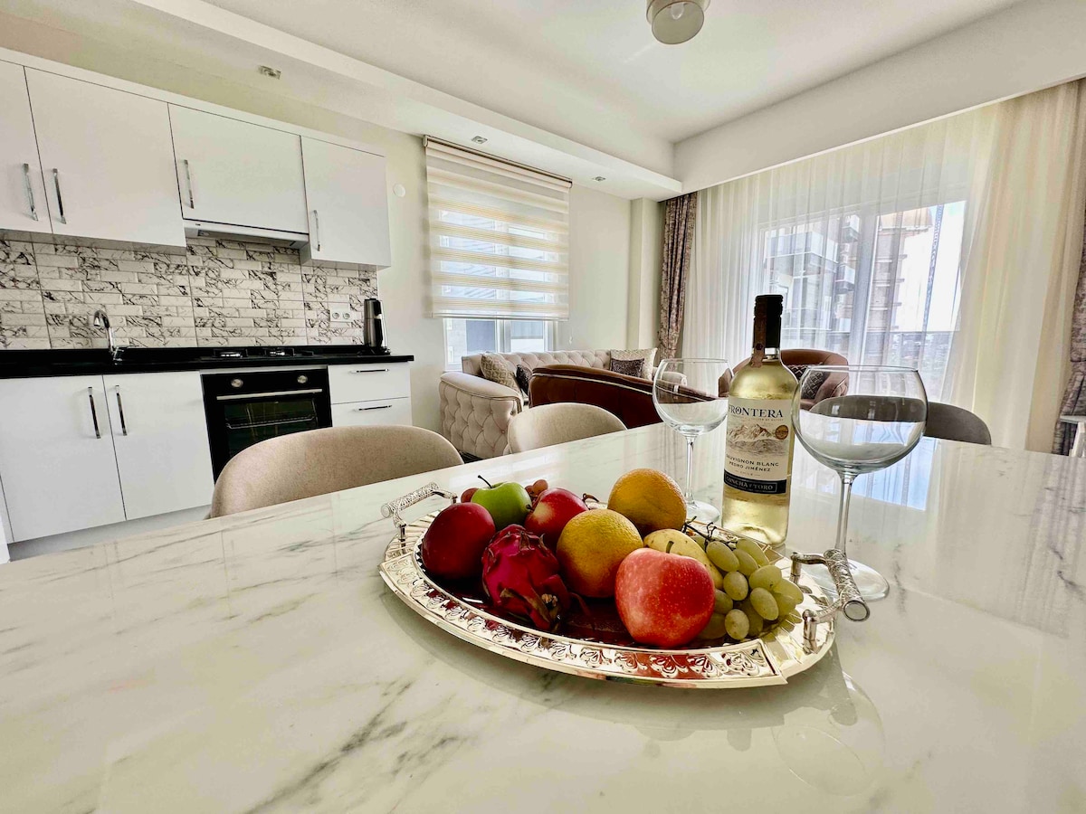 A modern kitchen is visible, featuring sleek cabinetry and a countertop with a decorative silver platter. Fresh fruits and a bottle of wine are arranged on the platter, with two glasses positioned alongside. Light pours in from large windows, enhancing the inviting space.