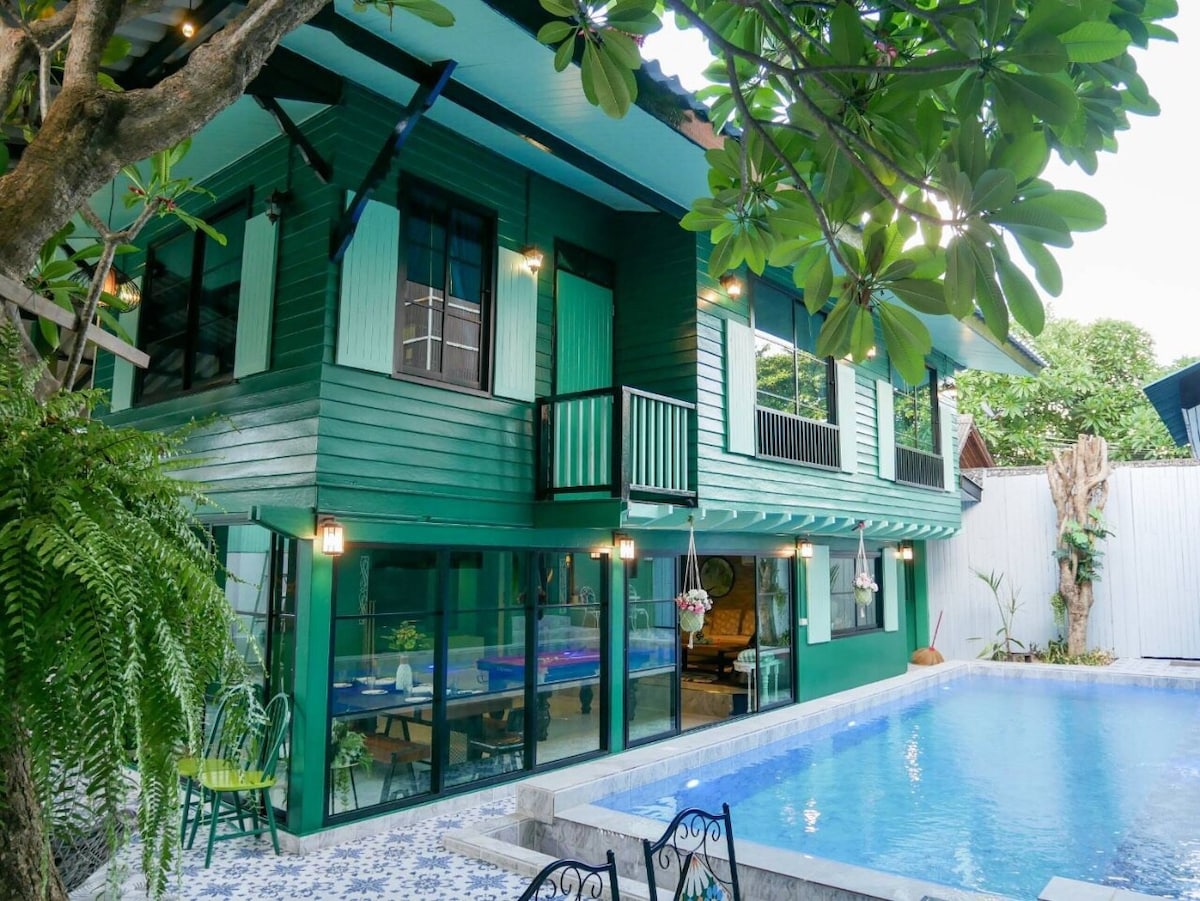 A two-story green villa is showcased with large glass windows, allowing natural light to fill the interior. A refreshing swimming pool is set in the foreground, surrounded by contemporary outdoor furniture and lush greenery, creating a serene environment.