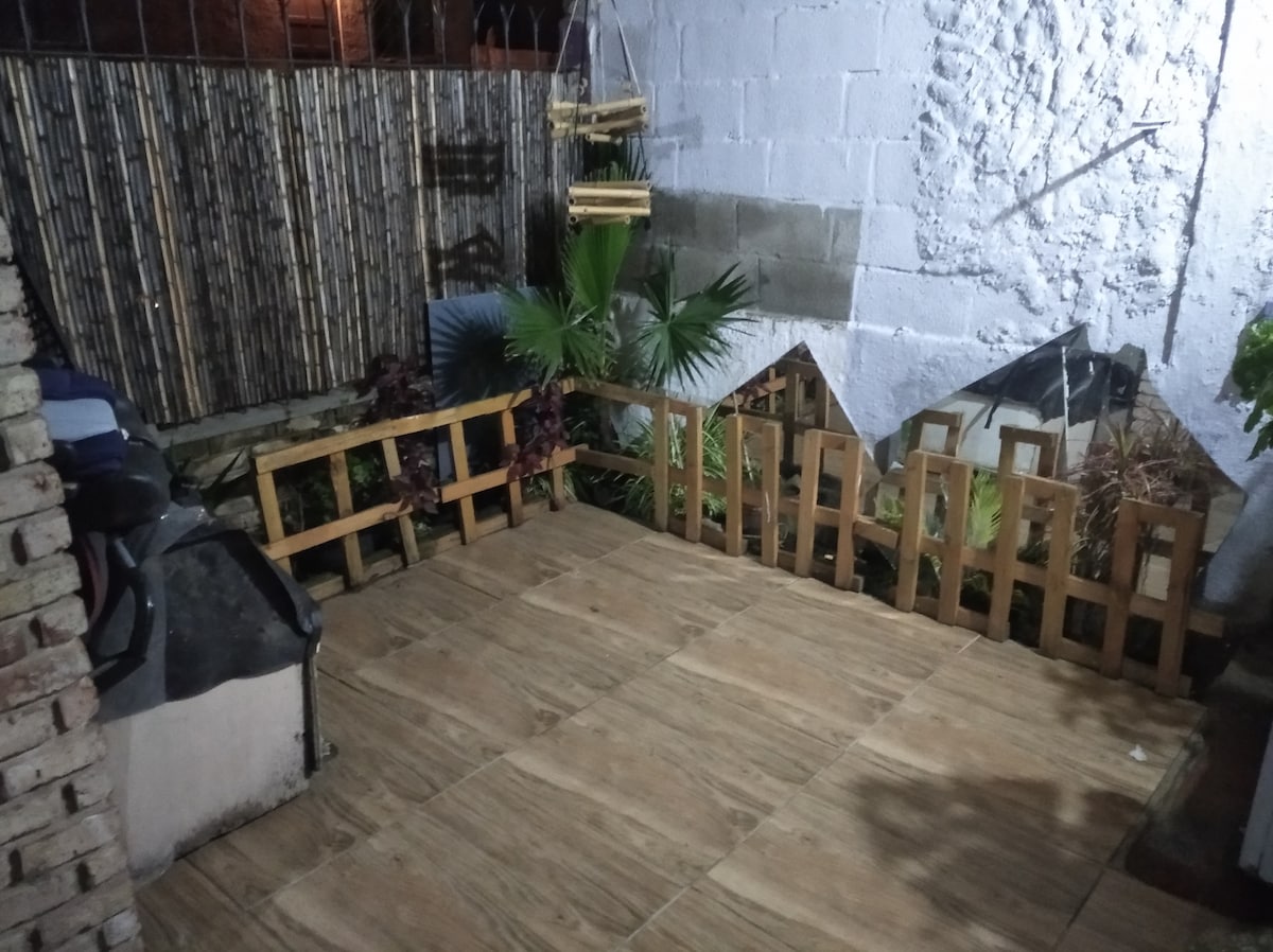 An outdoor area features a tiled flooring and a wooden fence enclosing a small garden. Green plants, including a palm, are integrated into the space. Soft lighting casts a gentle glow, enhancing the tranquility of this private retreat.