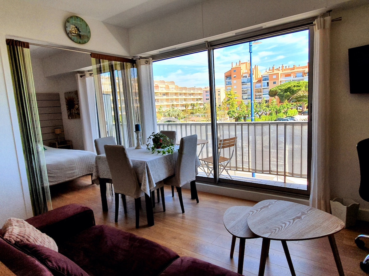 A spacious living area features large glass doors that open to a balcony, offering views of the surrounding landscape. A dining table with chairs and a small coffee table are positioned within the room, while soft natural light brightens the interior.