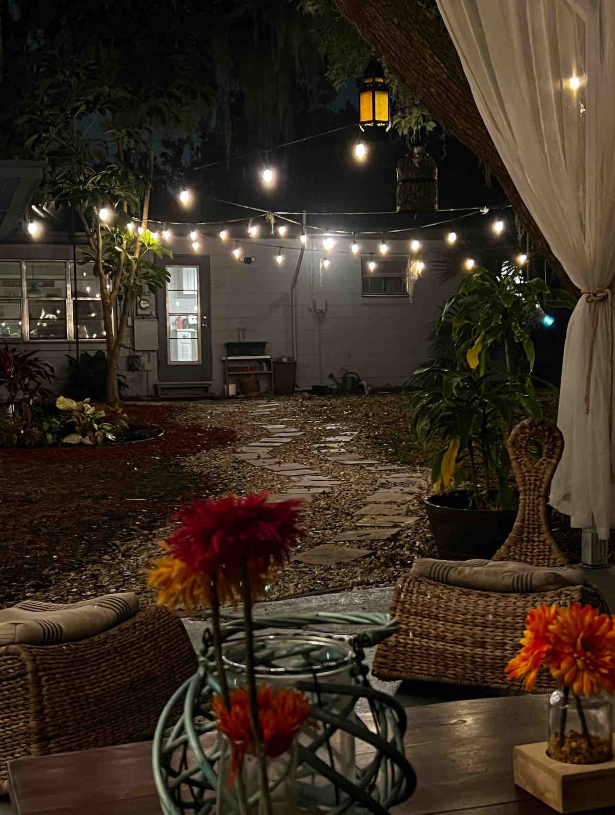 An outdoor area is highlighted by soft lighting from hanging string bulbs, creating a serene ambiance. A stone pathway leads through lush greenery, flanked by potted plants. Woven seating arrangements with decorative flowers are positioned nearby, inviting relaxation in an intimate setting.