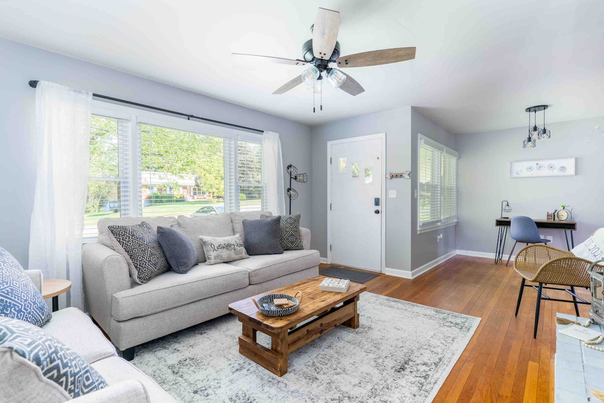 A spacious living room features a light gray sofa adorned with patterned cushions, situated on a soft area rug. Natural light fills the space through large windows with sheer curtains. A wooden coffee table and an accent chair provide additional seating, while a small dining area is visible in the background.