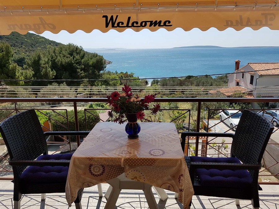 A balcony with a scenic view of the sea is displayed, featuring a round table set for two with a floral tablecloth. Two black wicker chairs flank the table. A welcoming sign is visible on the awning above, with greenery and the coastline visible in the background.