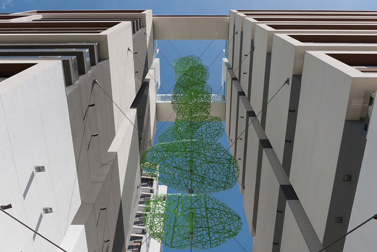 A modern building's design is highlighted, featuring an open air atrium with a large green sculpture suspended above. The structure showcases vertical lines and a mix of textures in its façade, with clear blue skies visible overhead.
