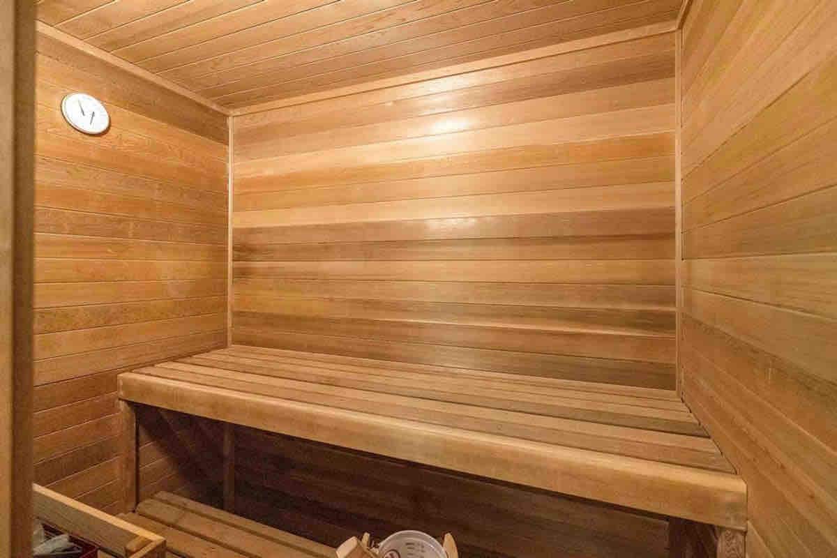 A warm wooden sauna interior is depicted, featuring smooth, horizontal plank walls and a spacious bench along one side. A small clock is mounted on the wall, contributing to the serene environment created by the natural wood tones.