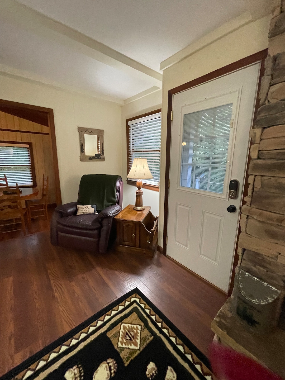 A cozy entryway features a comfortable armchair next to a wooden side table with a lamp. Natural light enters through a nearby window, while the stone fireplace adds a touch of character to the space. A welcome mat lies on the floor.