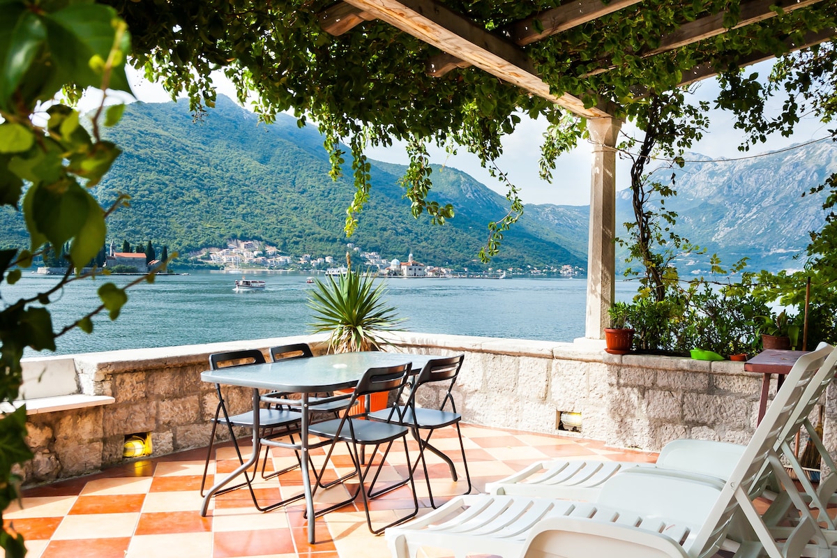 A spacious terrace is seen with a dining table surrounded by chairs, offering a clear view of the Bay of Kotor. Lush greenery drapes overhead, enhancing the outdoor dining experience. The tranquil waters and distant mountains form a picturesque backdrop against the stone wall.