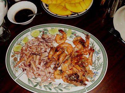 A large decorative plate is filled with an assortment of seafood, including grilled shrimp and calamari. Lime slices are placed around the dish, and a small bowl of dipping sauce is visible nearby. A platter of sliced mango sits in the background.