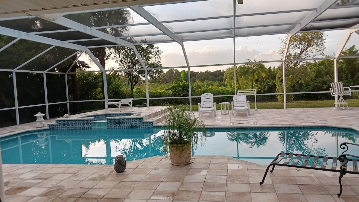 A screened-in pool area features a clear blue pool surrounded by a tiled deck. Several white lounge chairs are positioned around the pool, with a potted plant adding a touch of greenery. Lush landscaping is visible in the background, offering a serene environment.