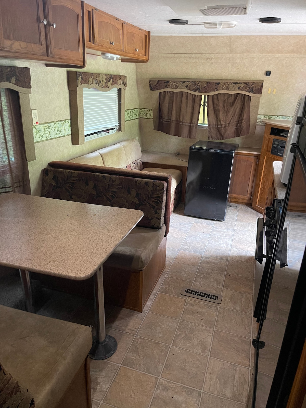 The interior of the RV features a cozy dining area with a table and booth seating. A compact refrigerator stands near the kitchen, with wood cabinetry above and a countertop visible. Light filters through partially closed curtains, highlighting the warm tones of the decor.
