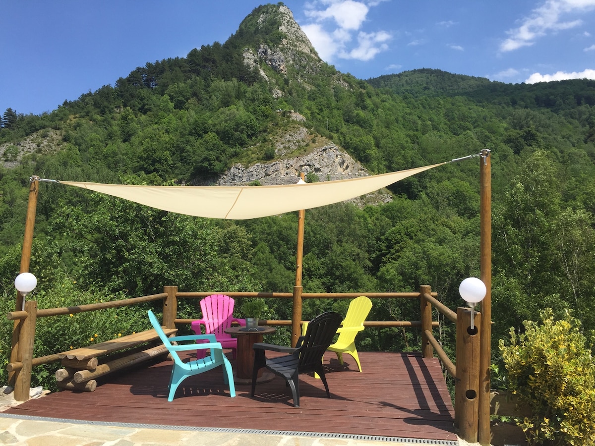 A wooden terrace is surrounded by lush greenery and mountainous scenery. Colorful chairs in pink, yellow, and black are arranged around a small table under a fabric shade, providing a relaxing outdoor space. Soft lighting fixtures are positioned for evening use.